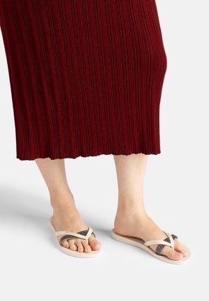 Barfuß in beigen Flip-Flops mit schwarzen, strukturierten Riemen, stehend auf einer hellen Oberfläche, mit einem teilweise sichtbaren roten, gerippten Rock.