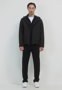 Giacca nera con zip e cappuccio, indossata sopra una maglietta nera, abbinata a pantaloni neri e sneakers bianche. Texture liscia, design minimalista.