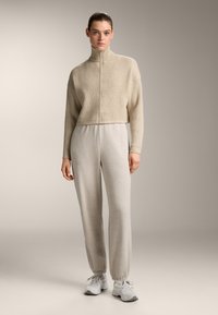 Beige Rippstrick-Zip-Pullover in cropped Länge, kombiniert mit hellgrauen Sweatpants mit elastischen Bündchen. Das Modell trägt hellgraue Sportschuhe.