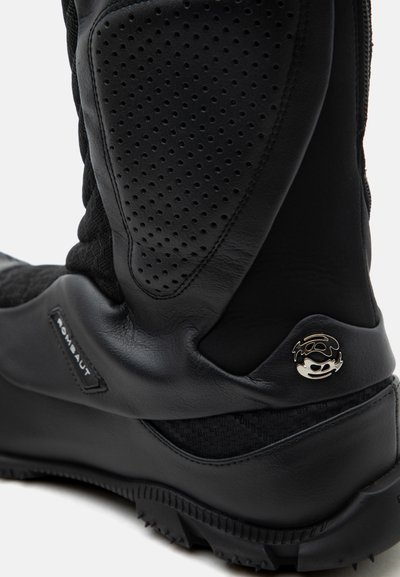 Κοντινό πλάνο μαύρης δερμάτινης μπότες με διάτρητες λεπτομέρειες, υφασμένη σόλα και μεταλλικό λογότυπο κοντά στη φτέρνα.