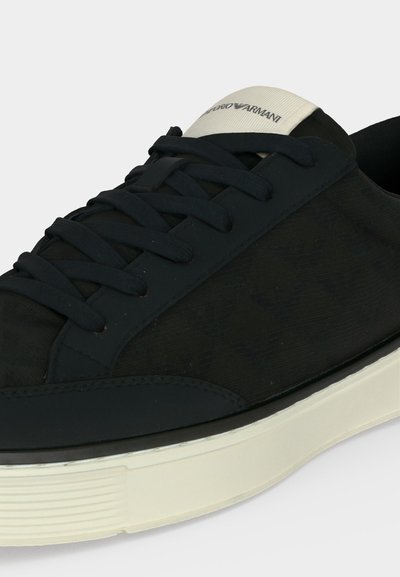 Baskets basses noires avec lacets noirs, semelle blanche et une étiquette blanche sur la languette affichant le logo Emporio Armani.