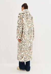 Hosszú, plüss kabát, amely műszőrme anyagból készült, krém és fekete foltos mintával. Magas gallérral, széles ujjakkal és térd alá érő fazonnal.