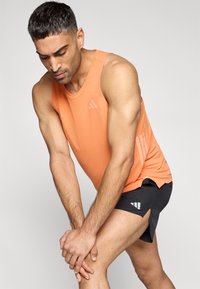 Πορτοκαλί αθλητικό τοπ με στρογγυλή λαιμόκοψη· διαθέτει διακριτικό λογότυπο και πλευρικές ρίγες. Συνδυάζεται με μαύρο σορτς που έχει πλαϊνό σκίσιμο.