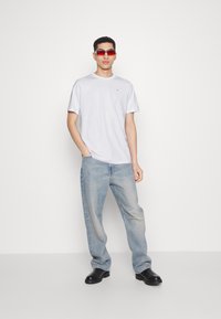 T-shirt bianca in cotone con un piccolo logo, jeans in denim blu chiaro e scarpe nere. Modello con occhiali da sole rossi, in piedi davanti a uno sfondo semplice.