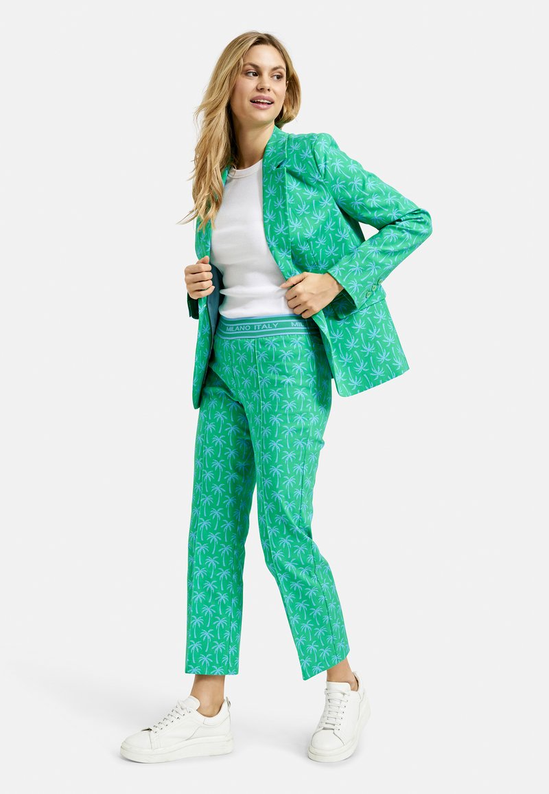 Green palm-patterned suit made of lightweight fabric; tailored jacket over fitted pants. White sneakers complete the look.