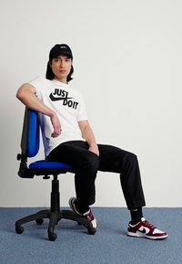 Hvid t-shirt med "JUST DO IT" tryk, sorte bukser, siddende på en blå kontorstol, iført en sort kasket og bordeaux sneakers.