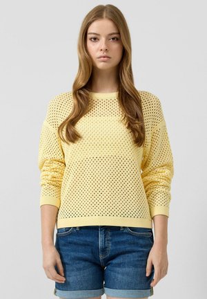 Młoda kobieta z długimi falującymi włosami, ubrana w żółty sweter o otwartym splocie i podwinięte krótkie jeansy, stojąca na jednolitym tle.