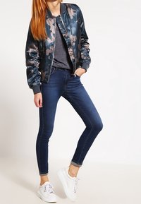 Srebrno-navy kamuflażowa kurtka bomberka z prążkowanym kołnierzem, połączona z ciemnymi dopasowanymi dżinsami i białymi sneakersami. Gładki materiał, dopasowany krój.