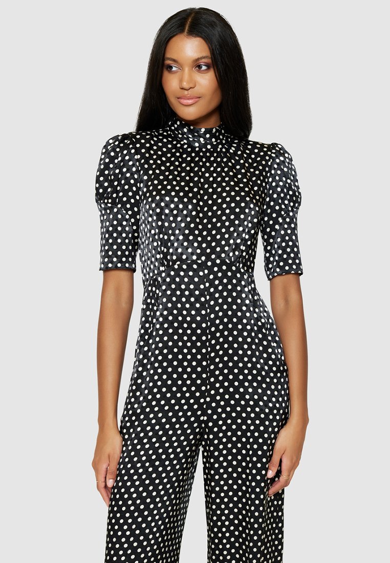 Czarny kombinezon w białe kropki, z wysokim dekoltem, krótkimi bufkami i dopasowaną górą. Gładka tekstura, szerokie nogawki.