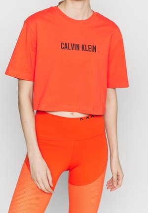 Mujer que lleva una camiseta corta de Calvin Klein de color naranja brillante y leggings de talle alto con paneles de malla, de pie frente a un fondo claro.