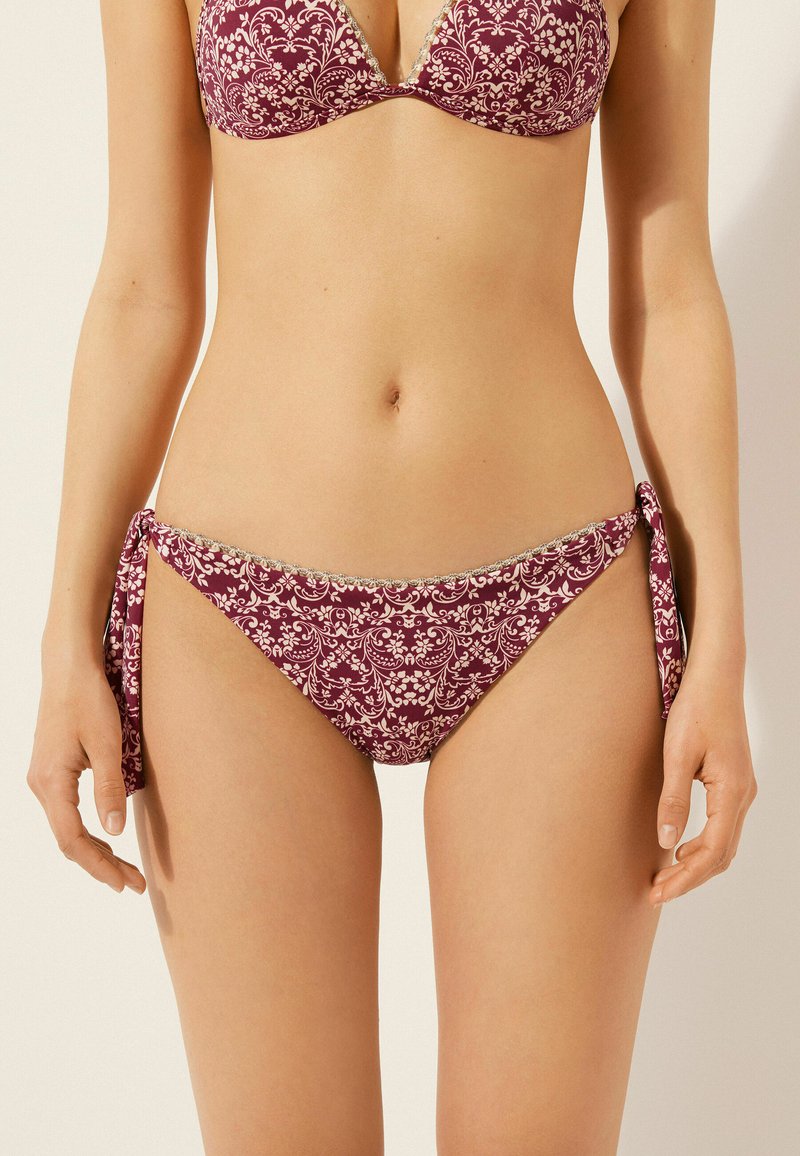 Bikini de un burdeos oscuro con intrincados patrones florales; parte superior triangular con tirantes ajustables y braguitas de bikini con lazo a los lados y con borde de encaje.