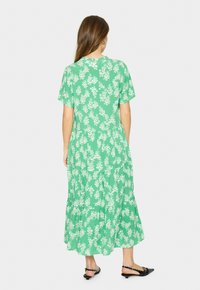 Rochie maxi floral verde cu mâneci scurte, design în straturi și model cu flori albe. Prezintă o croială relaxată și un material ușor. Pantofi plate negri.