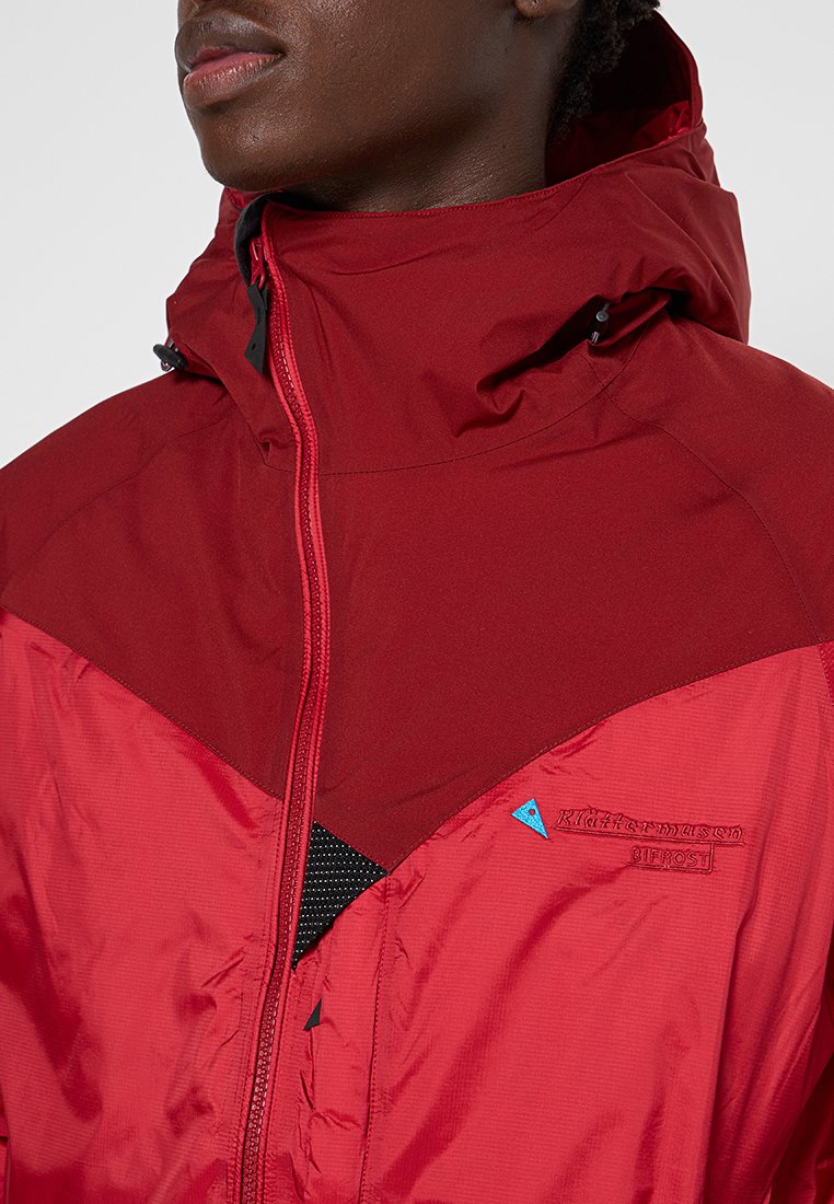 Röd vattentät jacka med dragkedjestängning, justerbar huva, nätaccent och broderad logotyp. Texturerad tyg med färgblockdesign.