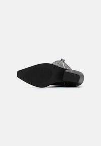Black heeled shoe with a smooth sole, featuring a textured bottom for grip, and a transparent strap across the front.