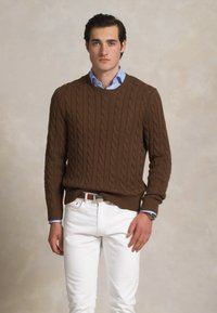 Pull en maille torsadée marron avec un col rond, porté par-dessus une chemise à col bleu clair et associé à un pantalon blanc. Montre visible au poignet.