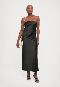 Vestido maxi de satén negro con un diseño sin tirantes, que presenta una textura suave y un ligero drapeado. Combinado con unos tacones negros con tiras y un bolso crema.