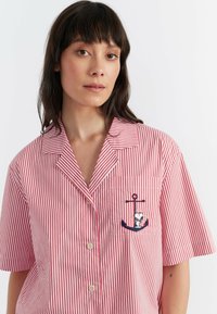 Chemise à manches courtes présentant des rayures verticales rouges et blanches, un col à encoches, et un design de ancre brodé sur la poche de poitrine.