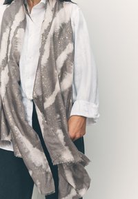 Sciarpa con motivo grigio e bianco con accenti dorati metallici, realizzata in un tessuto morbido e strutturato con bordi sfrangiati, indossata sopra una camicia bianca.
