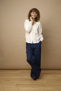 Femme aux cheveux bouclés sourit, portant un chemisier blanc brodé et un pantalon large bleu foncé, se tenant sur un sol en bois contre un mur beige.