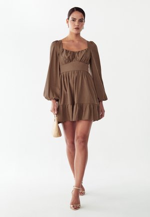 Kvinde iført en brun minikjole med lange ærmer, der holder en lille flet håndtaske og går fremad mod en ensfarvet hvid baggrund.