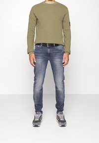 Olivgrön långärmad skjorta, blå jeans, svart bälte, grå sneakers med gröna accenter. Avslappnad passform, texturerad tyg, standard denimskärning.