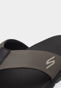 Κοντινό πλάνο μαύρου σανδαλιού τύπου slide με υφασμένη γκρι λουρίδα που διαθέτει μικρό μοτίβο από τελείες και μεταλλικό λογότυπο "S" στο πλάι.