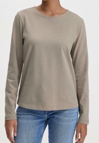 Taupe långärmad skjorta på person som bär blå jeans, visad från halsen till höfterna mot en enfärgad bakgrund.