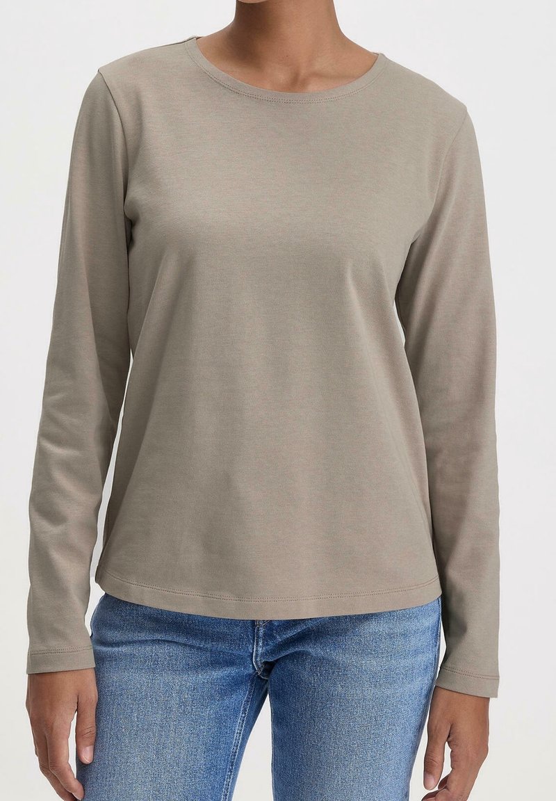 Taupe långärmad skjorta på person som bär blå jeans, visad från halsen till höfterna mot en enfärgad bakgrund.