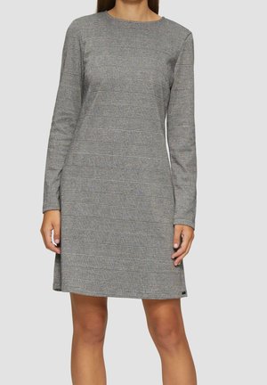 Femme portant une robe grise à carreaux, manches longues, longueur au genou, avec un col rond, debout devant un fond clair uni.