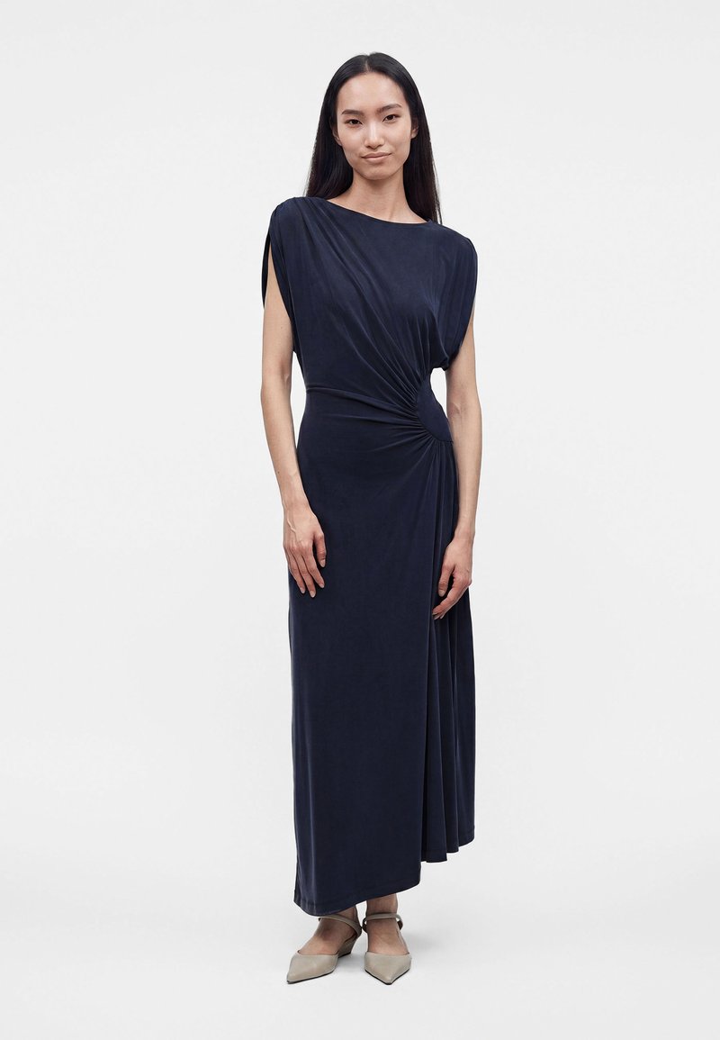 Femme debout dans une robe longue bleu marine sans manches, drapée avec un nœud sur le côté, chaussures pointues neutres et cheveux foncés raides.