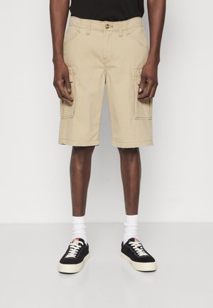 Mann trägt beige Cargo-Shorts, weiße Socken und schwarze Sneakers, steht vor einem schlichten weißen Hintergrund.