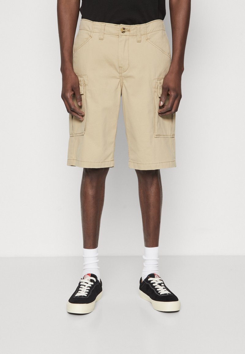 Homme portant un short cargo beige, des chaussettes blanches et des baskets noires, debout sur un fond blanc uni.