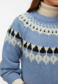 Ženska, ki nosi modri pleteni pulover s črno-belimi geometričnimi vzorci čez bež rebrasti puli.
