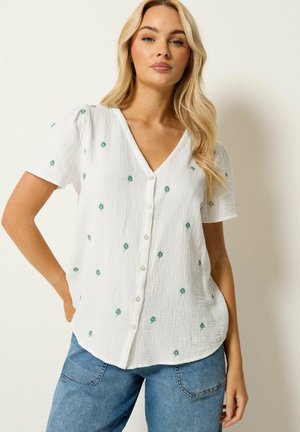 Frau mit langen blonden Haaren trägt eine weiße, kurzärmelige, geknöpfte Bluse mit kleinen grünen Blumenmustern und eine blaue Jeans, steht vor einer schlichten Wand.