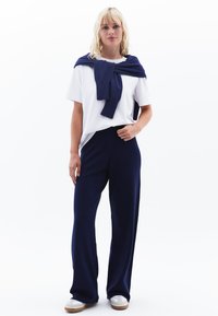 Witte t-shirt, marineblauwe losse broek en een marineblauwe trui die over de schouders is gedrapeerd. Schoenen zijn licht met een gestructureerde zool. Eenvoudig, casual ontwerp.