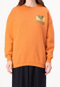 Oranssi huppari, jossa on rento malli, ribbaustakkiset hihat ja alareuna. Vasemmassa rinnassa on logo painatuksena vihreänä ja kermanvärisenä.