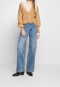 Hellbraune Strickjacke mit tiefem V-Ausschnitt, darunter weiße Spitzenborte, kombiniert mit hoch taillierten blauen Jeans mit weitem Bein und schwarzen Stiefeletten.