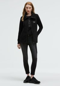 Black fleece jacket with zip closure, featuring a high collar and front pockets, paired with black slim jeans and black sneakers.