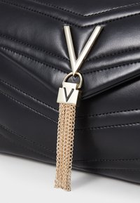 Black quilted leather handbag featuring a gold-tone 'V' hardware accent and a decorative chain tassel. Textured surface adds depth.