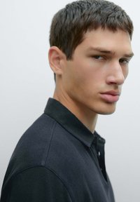 Svart polo skjorta med kort krage, med en slät textur och tre knappslå, bärs av en modell med kort hår.