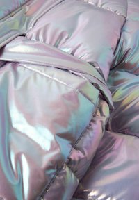 Veste en duvet irisé avec une surface brillante et réfléchissante, un design matelassé et un détail de ceinture nouée. Les couleurs varient entre le violet et l'argent.