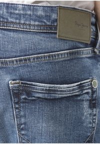 Džínové kalhoty střední modré barvy s opotřebovanou texturou. Mají koženou nášivku a zadní kapsu. Obsahují kovový knoflík na pase.