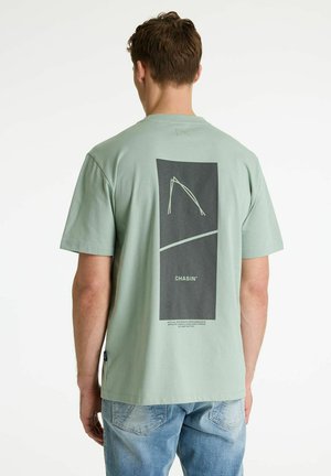 Mees, kes kannab helerohelist T-särki musta ristkülikukujulise graafika ja tagaküljel oleva tekstiga "CHASIN'", kombineeritud heleroheliste teksadega.