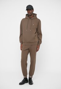 Brun sweatshirt med huva och framficka, kombinerad med matchande avsmalnande byxor. Har märkessymboler och kontrasterande muddar. Svart keps och skor.