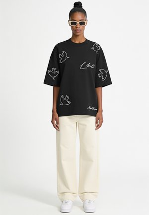 Femme portant un t-shirt noir oversize avec des dessins d'oiseaux blancs et un texte, un pantalon large crème, des baskets blanches et des lunettes de soleil crème.