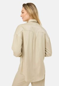 Chemise beige à manches longues avec une coupe décontractée, texture lisse, ourlet arrondi et col légèrement surdimensionné, présentant des détails de plis au dos.