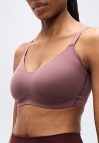 Αθλητικό σουτιέν Nike σε μωβ απόχρωση, κατασκευασμένο από λείο ύφασμα. Σχεδιασμός με V ντεκολτέ, λεπτές ράντες στους ώμους και διακριτική λεπτομέρεια με το λογότυπο κάτω από το ντεκολτέ.