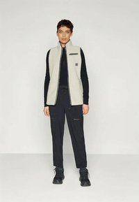 Hele hall fleece'i vest kõrge kaelusega ja eest lukuga, kannab musta pikakäiste särgi peal ning on kombineeritud mustade cargo-pükste ja saappastega.