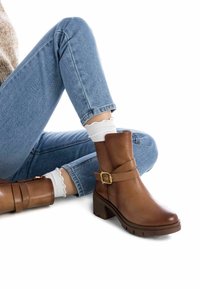 Botas de cuero marrón hasta el tobillo con un detalle de correa y hebilla, puntera cuadrada y tacón grueso. Combinadas con jeans de color azul claro y calcetines blancos con volantes.