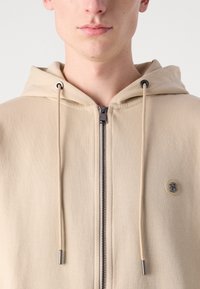 Felpa beige con zip realizzata in un tessuto morbido, dotata di una zip argento e lacci con regolazione. Presenta un piccolo logo sul petto.
